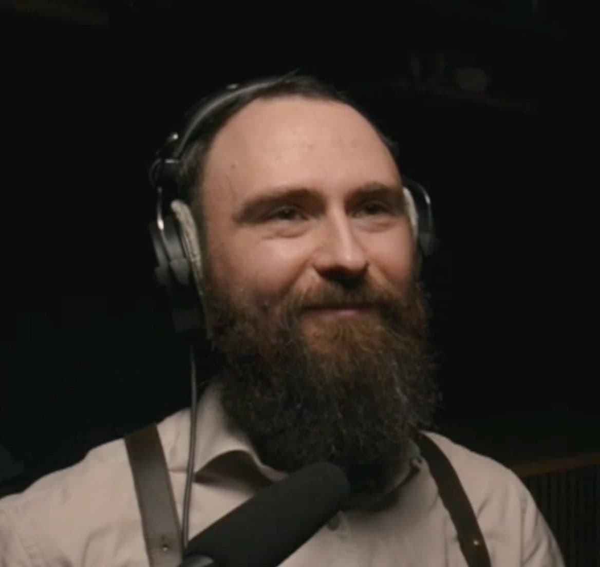 Anton Malinski, sporting a full beard and headphones, smiles while discussing Testing Pipelines into a microphone, dressed in a light-colored shirt and suspenders against a dark background.